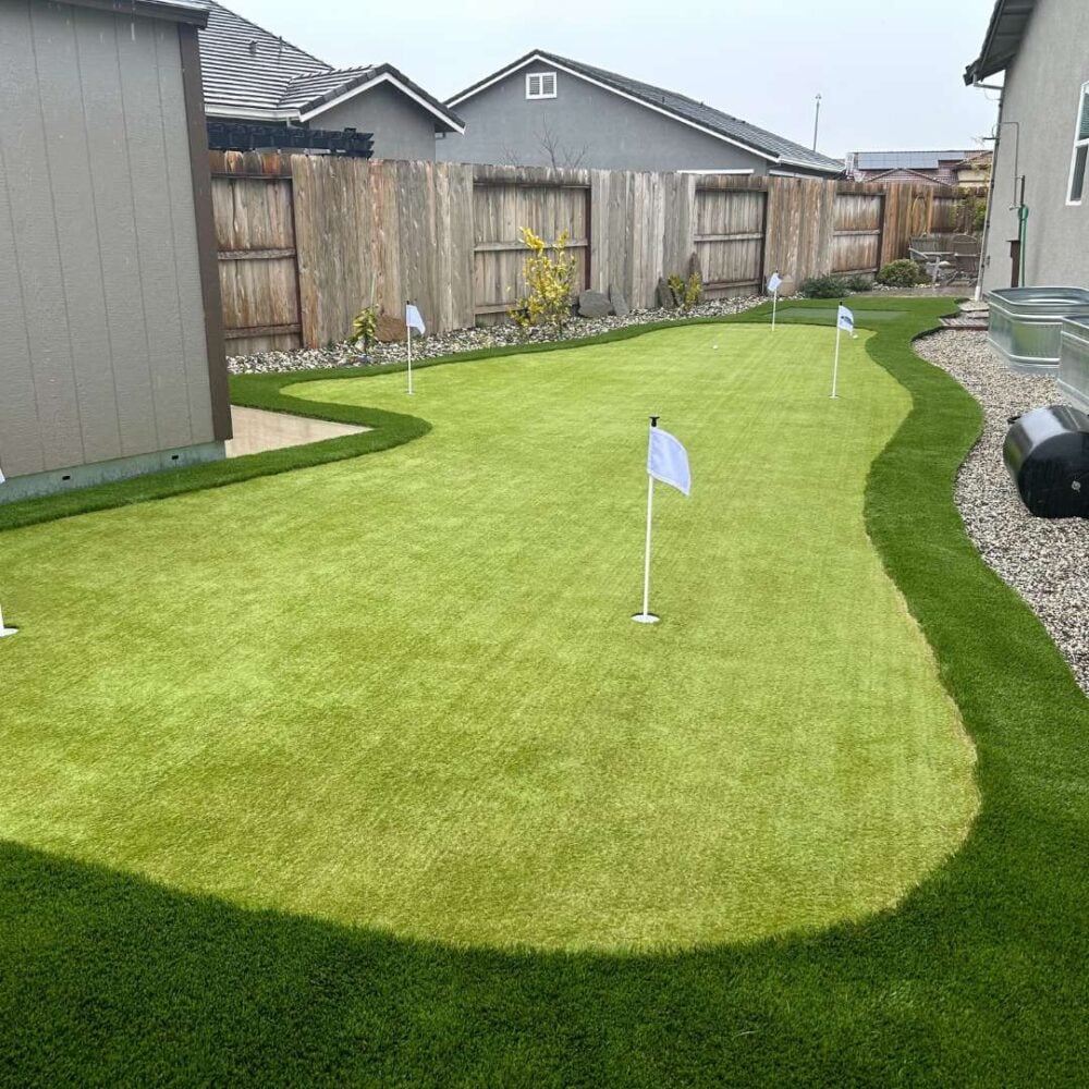 Backyard putting green installation featuring multiple white flags on lush, artificial turf, surrounded by a landscaped area with gravel and fencing, showcasing a premium artificial turf solution by Heavenly Greens.