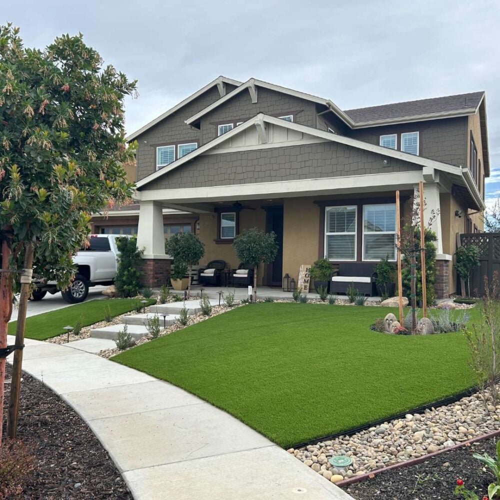Front yard featuring lush artificial turf installation, surrounded by landscaped plants and a modern home with a porch, showcasing low-maintenance landscaping solutions by Heavenly Greens.