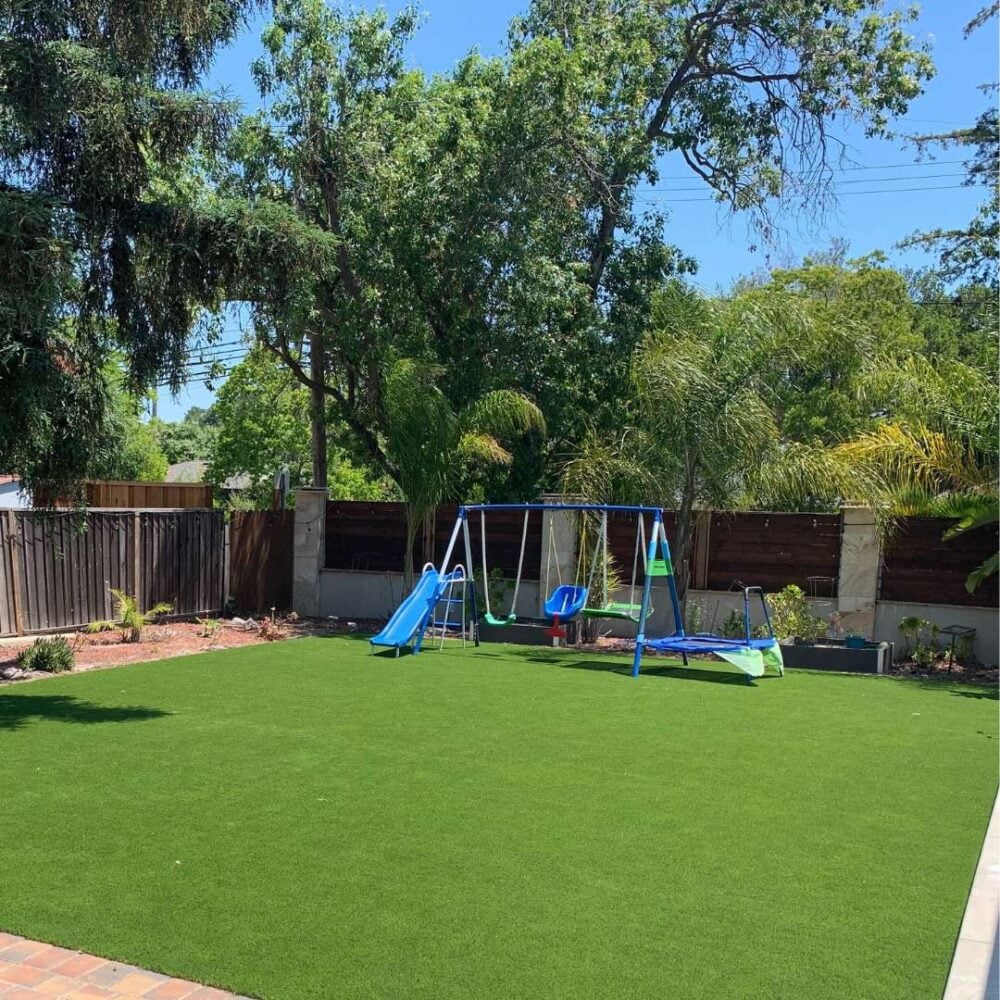 Kid's play area featuring vibrant artificial turf, swing set, and slide, surrounded by trees and landscaping, showcasing Heavenly Greens' family-friendly outdoor solutions.