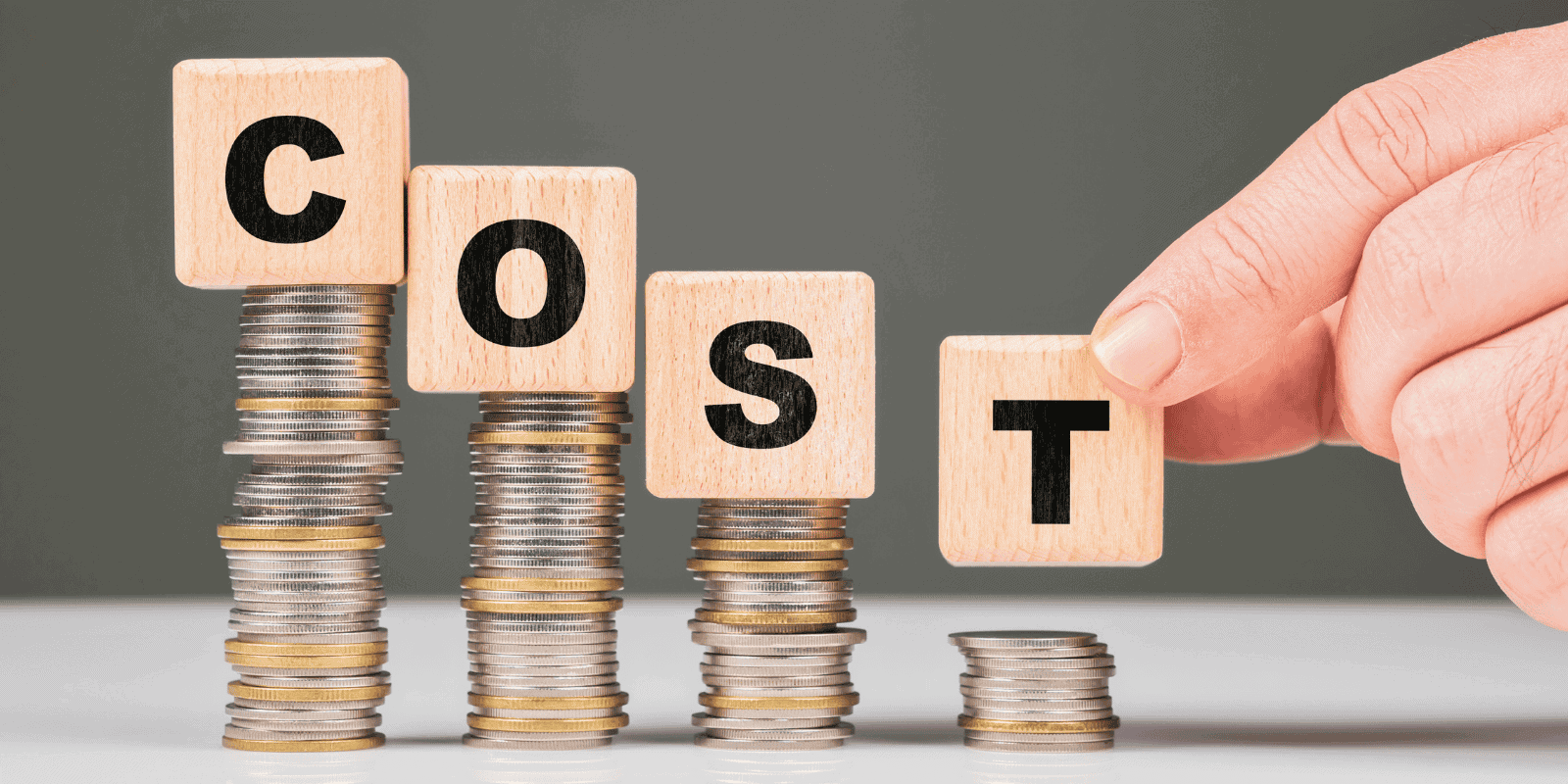 Hand placing wooden block with letter "T" atop stacks of coins spelling "COST," symbolizing expenses related to artificial grass installation.