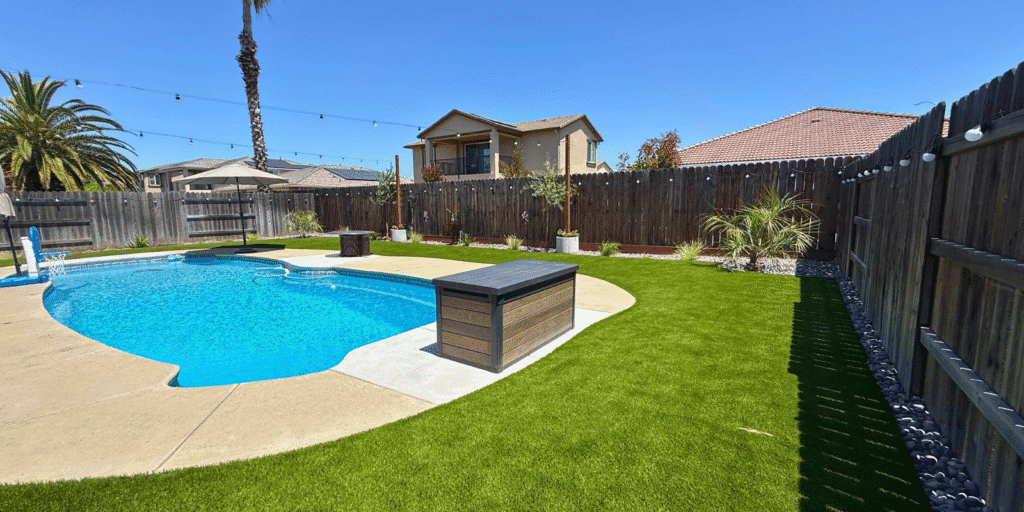 Backyard with artificial grass surrounding a swimming pool, featuring palm trees, a wooden fence, and a stylish outdoor storage box, illustrating a modern outdoor living space.
