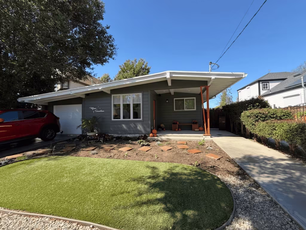 Residential property featuring a well-maintained artificial grass front yard, surrounded by landscaping elements, with a modern grey house and a red vehicle parked nearby, illustrating sustainable lawn solutions by Heavenly Greens.