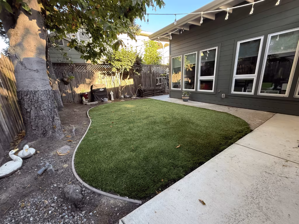 Backyard with lush artificial grass installation, surrounded by a tree and patio area, showcasing a well-maintained outdoor space ideal for homeowners seeking sustainable lawn solutions.