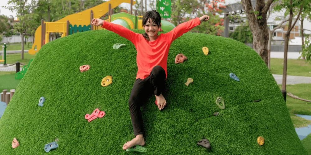 Child playing on artificial grass climbing structure in a park, showcasing fun and safe recreational area design with colorful climbing holds.