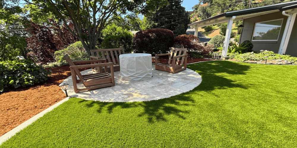 Outdoor seating area featuring artificial grass surrounding a circular stone patio with wooden chairs and a table, framed by lush greenery and garden landscaping, illustrating modern outdoor living design.