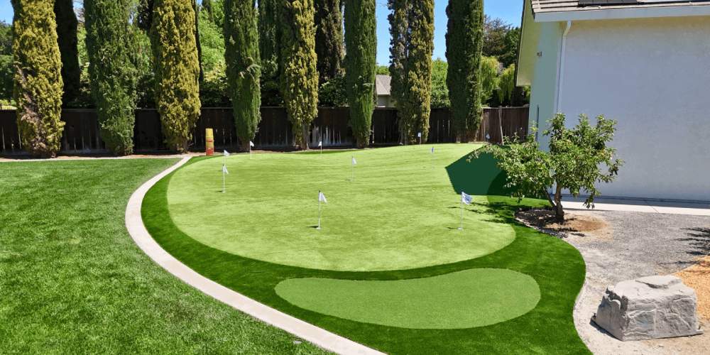 Artificial grass golf putting green with multiple flags, surrounded by lush greenery and a residential backyard setting, illustrating convenient home golf practice area.