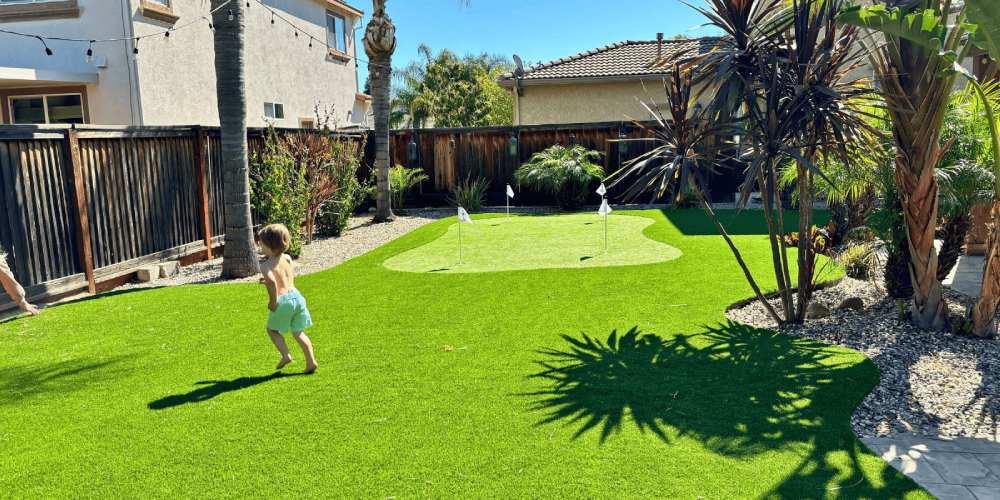 Child playing on lush artificial grass lawn with putting green in a sunny backyard, featuring palm trees and decorative landscaping, showcasing the benefits of synthetic turf for outdoor spaces.