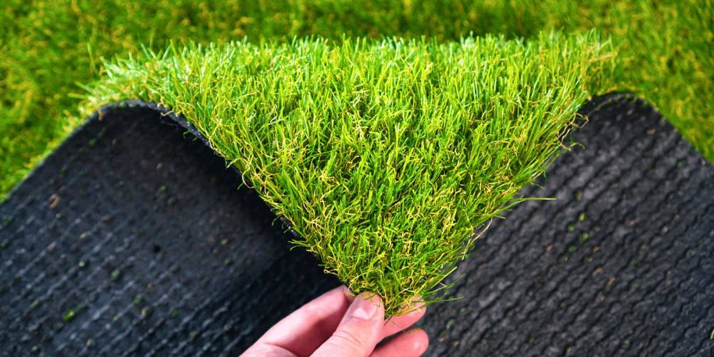 Hand holding a piece of artificial grass showcasing lush green blades and black backing, illustrating options for backyard turf solutions.