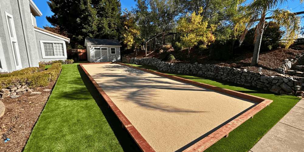 Bocce ball court surrounded by artificial grass and landscaped yard, showcasing a comfortable outdoor space for gatherings and leisure activities.